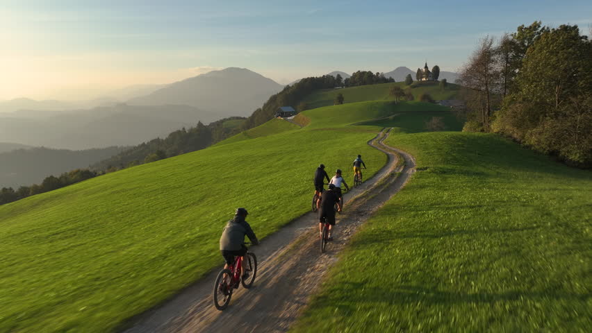 AERIAL: Mountain bikers ride along the hilltop trail on a sunny autumn evening. Scenic drone shot of a group of active tourists riding electric bikes at sunrise. Bike ride in the Slovenian mountains.