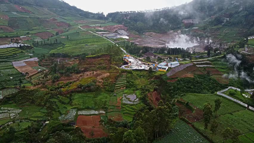 Aerial view of a mountainous landscape with terraced fields, a winding road, and a small settlement. Steam rises from geothermal activity in the background, indicating volcanic features.