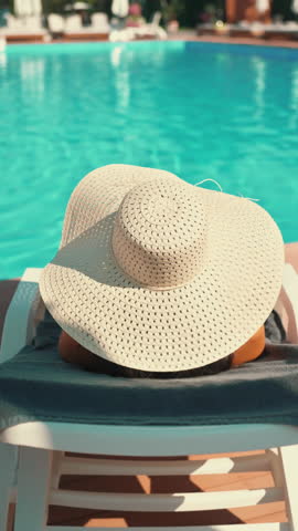 Vertical Screen: Woman reclines by pool, hands resting on large woven sun hat, summer relaxation. Vibrant turquoise water adds to tranquil atmosphere. Concept of vacation, leisure and sunbathing