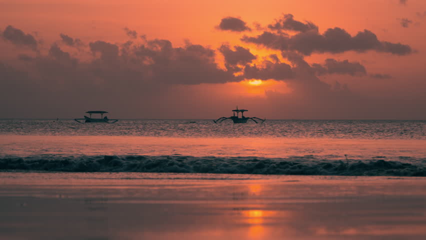 Couple Walks Barefoot in Shallow Sea Water At Bali Sunset - Outrigger Old Boat in Silhouette Anchored Floating In Ocean in Background. Bright Sun Behind Clouds, Indonesia