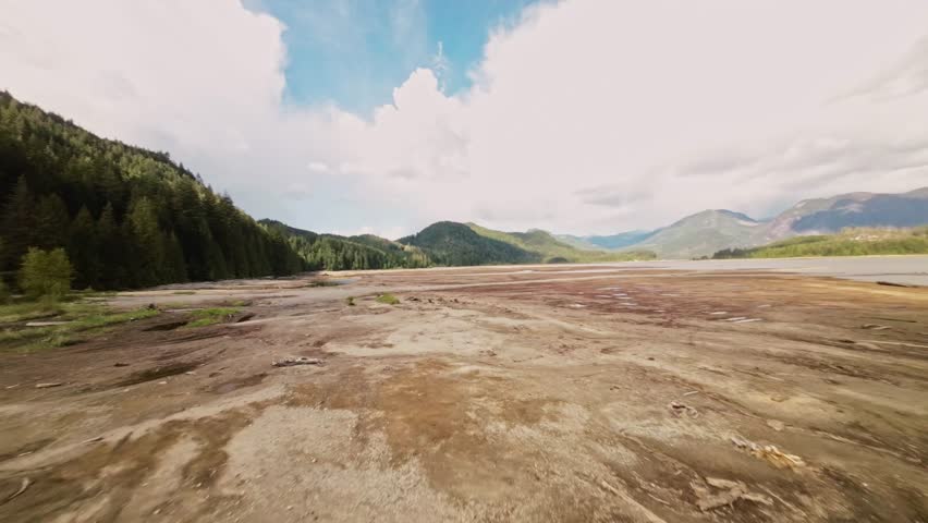 Low FPV aerial over dried-up expanse Stave Lake showcasing rugged rocks dust-laden fallen logs, tree stumps bright sunny day with wispy clouds in the sky Beautiful exciting adventurous terrain