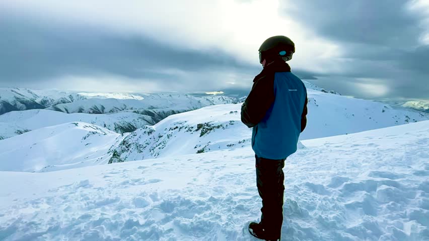 A man in snow clothes stands on a snowy summit and drinks from a can, wide shot in slow motion