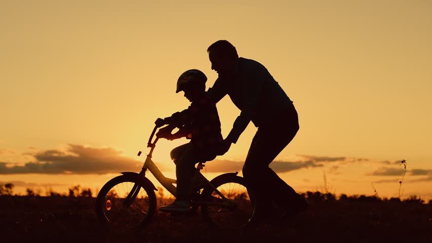 Child dream learns to ride bicycle. Father helps his daughter ride bike. Father teaches child wearing safety helmet to ride bicycle in park. Child rides bicycle. Kid, dad play together, sunset. Family