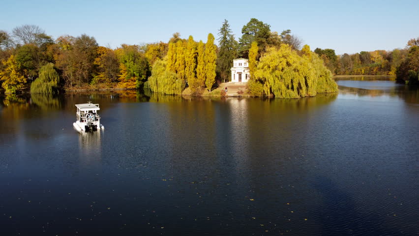Flying over surface of lake with a floating white boat. Many trees with yellow and green leaves and white architectural building on shores of lake on sunny autumn day. People walking in the park