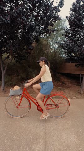 Side view of a woman riding her vintage red bicycle, surrounded by trees under a cloudy sky.