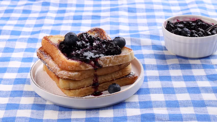 A white plate with garnished French toast on a blue napkin on the table. A woman’s hands cut the toast with blueberry jam using a knife and fork.