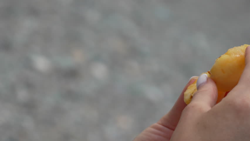 Close-up of hands holding a peeled orange segment. The orange is juicy and has a bright yellow color. The background is blurred and shows a gray surface.