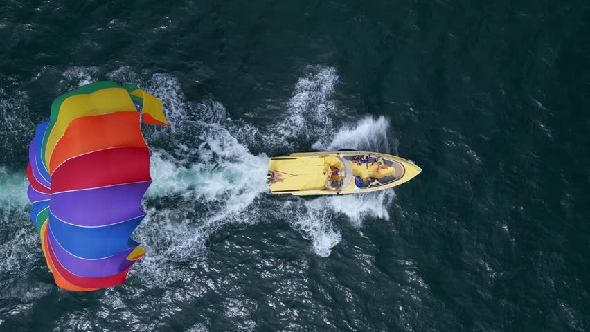 Drone video of a colorful parasail being towed by a speedboat over the ocean. The scene captures the excitement of parasailing with stunning views of the coastline and open sea