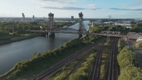 Aerial footage capturing a steel lift bridge over a river with adjacent railway tracks. Showcasing both natural greenery and urban skyline at sunrise - Powered by Shutterstock - Get 15% off with code: PIKWIZARD15