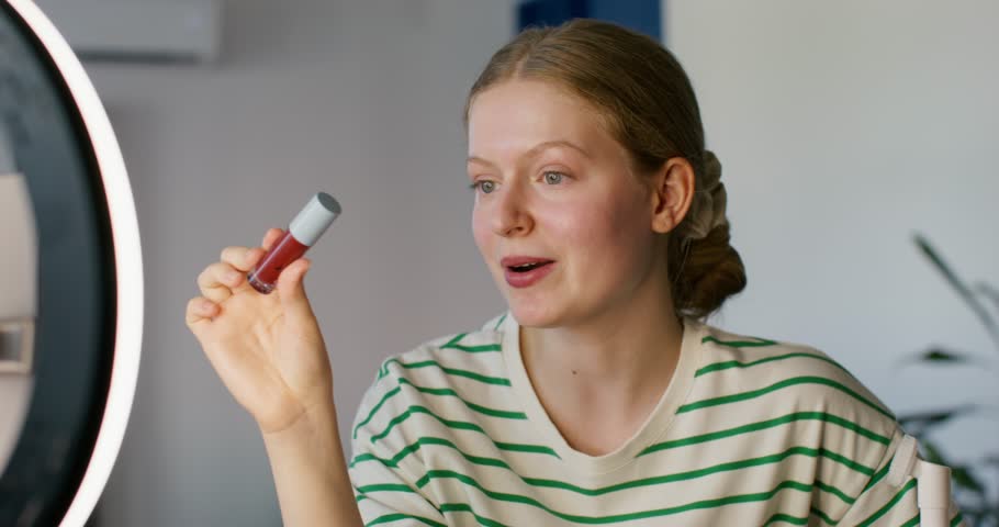 A young woman shoots a beauty blog on her mobile phone at home. She wears red lipstick and talks. A mobile phone and a round lamp on a tripod