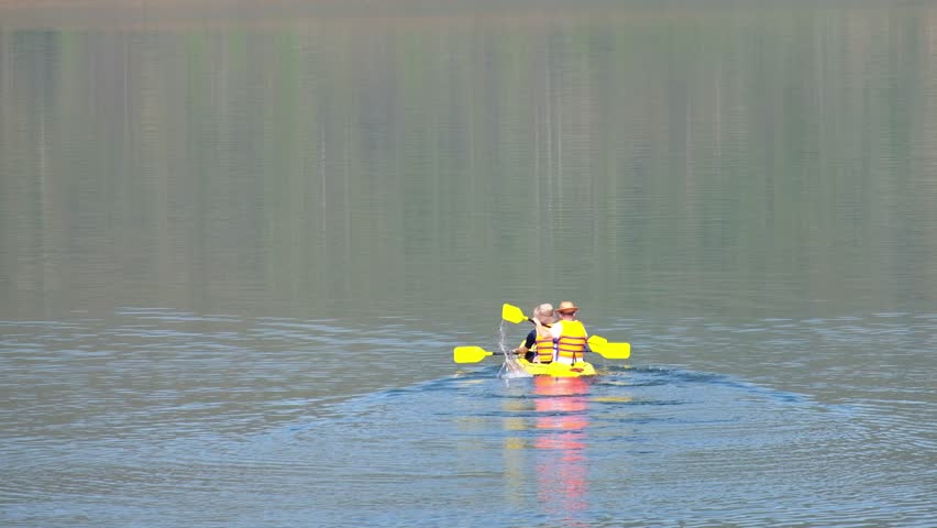 Two friends enjoy kayaking on a calm lake, paddling in sync with their bright yellow oars. The still water reflects their movements, creating a peaceful and enjoyable outdoor experience.