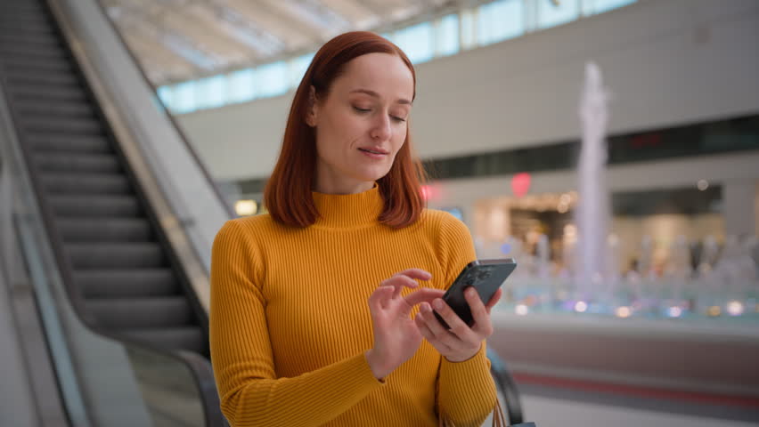 Young Caucasian girl female woman businesswoman client shopper buyer holding mobile phone cellphone using online store order app ordering searching lost credit card in shopping bags indoors shop mall