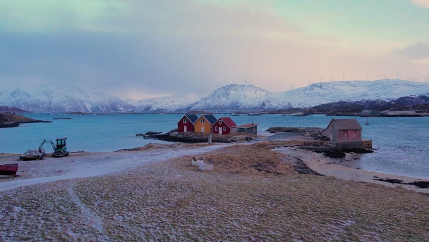 Aerial of small traditional rorbu fishing huts with snow covered mountain background, on winter Sommaroy island, Norway