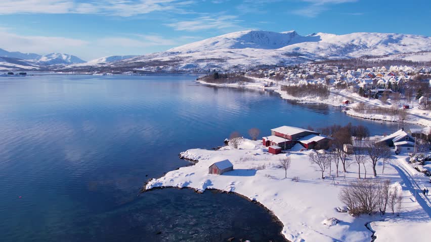 Snow covered mountain range on coastline in winter, Norway. Surroundings of town Tromso. Panoramic aerial view landscape of nordic snow cowered mountains, houses and ocean. Troms county, Fjordgard