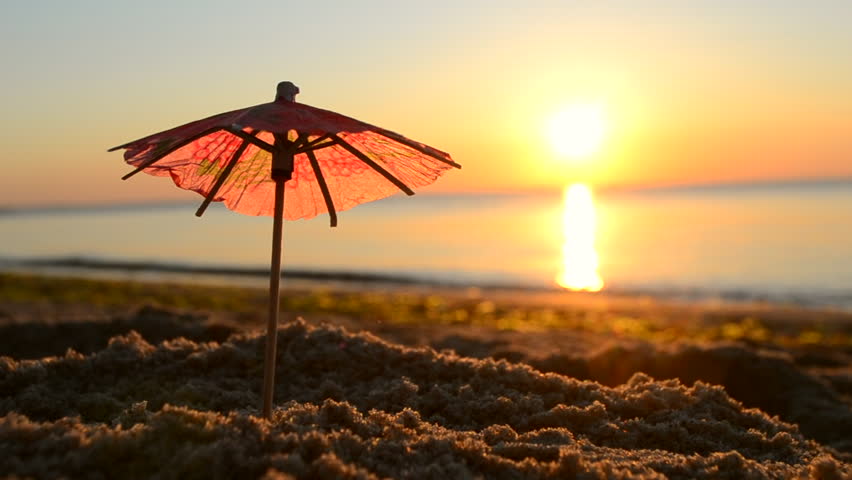 Paper cocktail umbrellas in sand on seashore at sunset dawn close-up. Beach umbrellas on seaside near sea ocean sunrise and sunset. One small umbrellas stand in beach sand background of sea waves