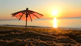Paper cocktail umbrellas in sand on seashore at sunset dawn close-up. Beach umbrellas on seaside near sea ocean sunrise and sunset. One small umbrellas stand in beach sand background of sea waves - Powered by Shutterstock - Get 15% off with code: PIKWIZARD15