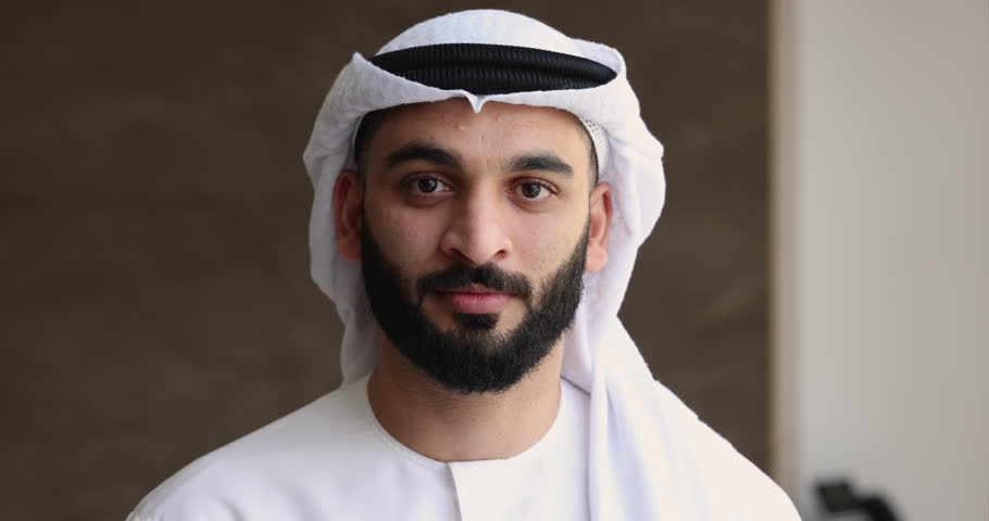 Serious handsome young Arabic businessman in white Muslim cloth and headwear posing for portrait, getting positive, cheerful, happy, looking at camera with toothy smile. Close up head shot