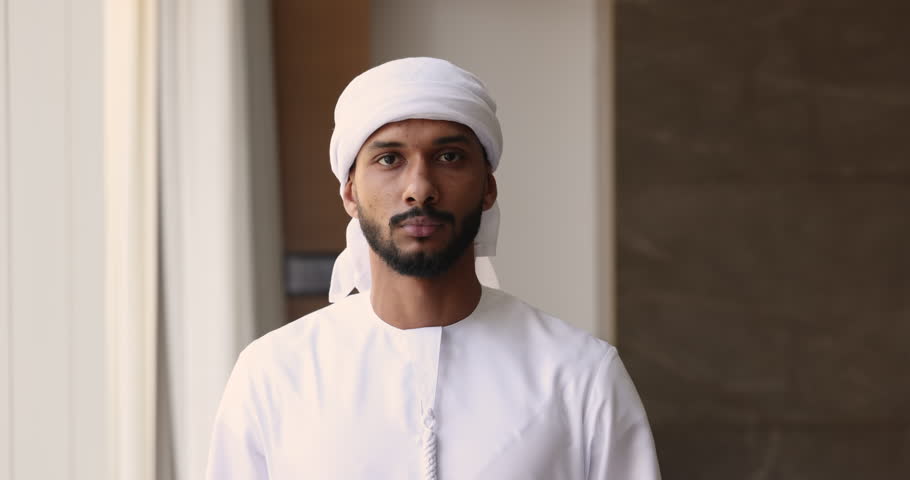 Serious handsome young Arabic business owner man in white Muslim clothes posing in office for front portrait, getting happy, looking at camera. Successful Arab businessman, boss, leader head shot