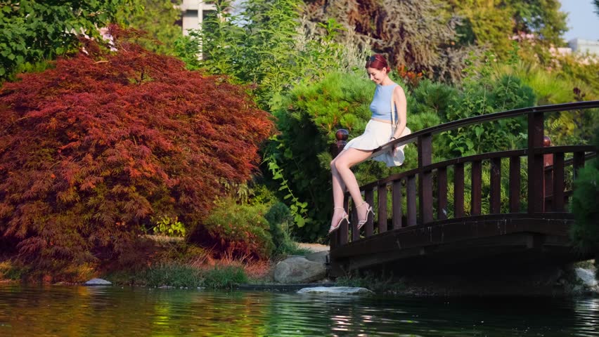 A young, beautiful red-haired woman wearing a white skirt and high heels is sitting on a bridge over a lake in a wooded forest, having fun with the water on a summer day