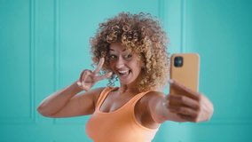 A cheerful curly woman makes video record selfie peace sign with her phone for social media in the studio on blue teal background - Powered by Shutterstock - Get 15% off with code: PIKWIZARD15