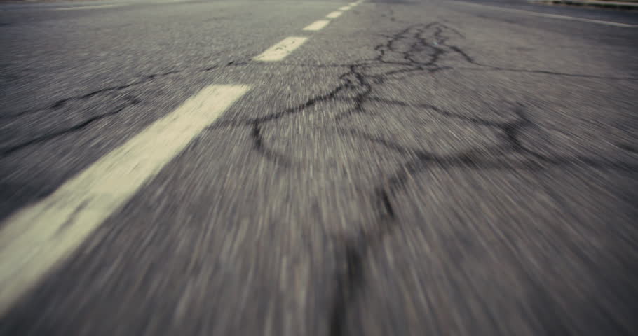 A low-angle, dynamic shot of a rural asphalt road at dusk, capturing the texture of the pavement as the vehicle swiftly moves forward, emphasizing the journey and the solitude of the empty street.