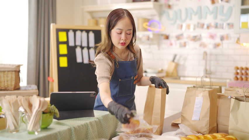 Portrait of Young Asian woman bakery shop owner preparing customer order bakery in delivery paper bag on kitchen counter. Small business e-commerce online catering food and drink entrepreneur concept.