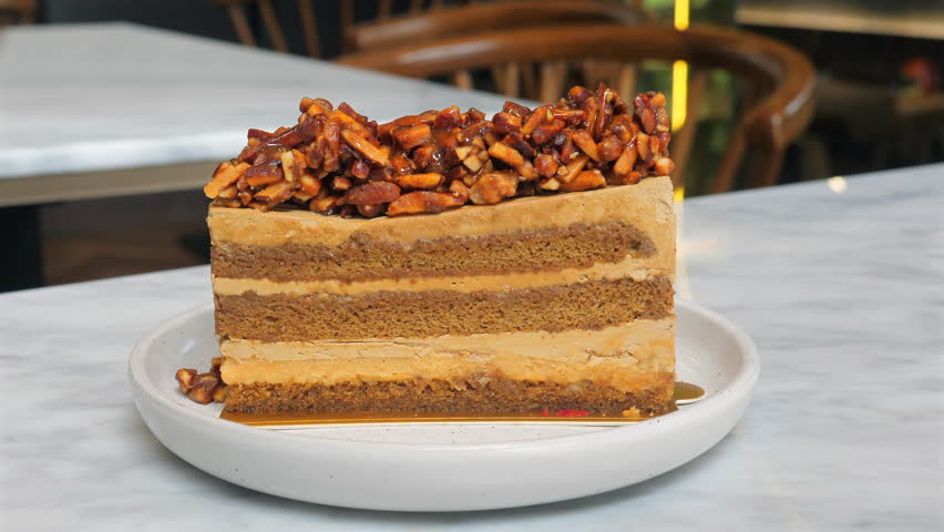 Slice of layered caramel cake topped with caramelized nuts on white plate, on marble table in cozy cafe, showcasing delicious dessert indulgence. Indulgence and Gourmet Treats.