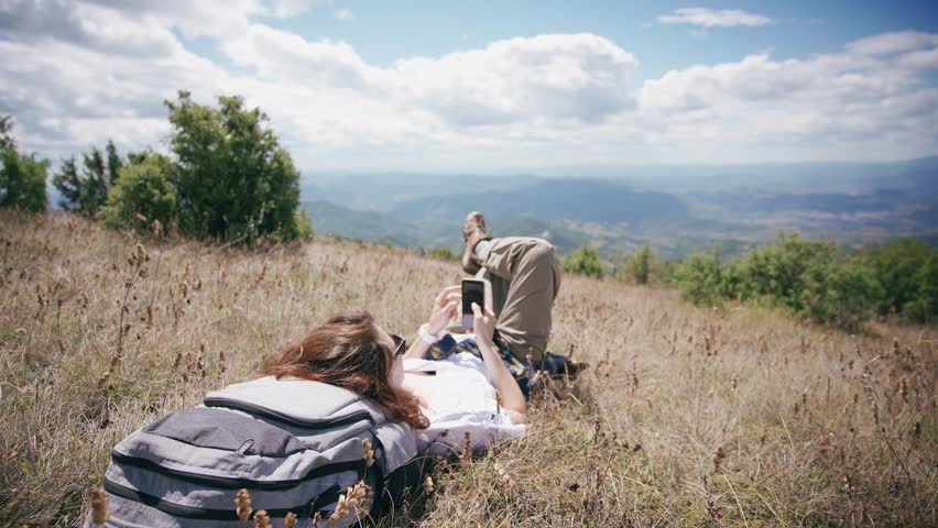 A young woman traveler using a smartphone while lying in the grass in the picturesque mountains.