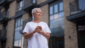 Low angle portrait of happy senior woman holding cup with iced coffee and looking up with smile on face, enjoying moment of relaxation outside on background of residential building, slow motion. - Powered by Shutterstock - Get 15% off with code: PIKWIZARD15