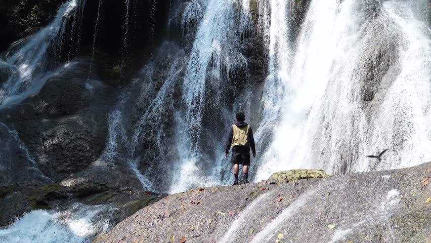 Young male tourist standing in front of steep stone wall and enjoying waterfall view. Environment, water circulation, tourism concept.