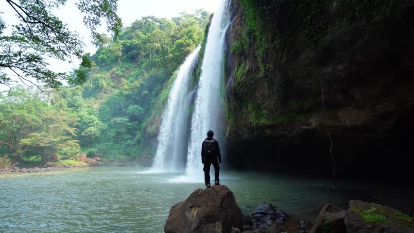 Man stands on a rock and raising her hands up in the air wiht beautiful Sodong Waterfall, Geopark Ciletuh, Sukabumi, Indonesia.