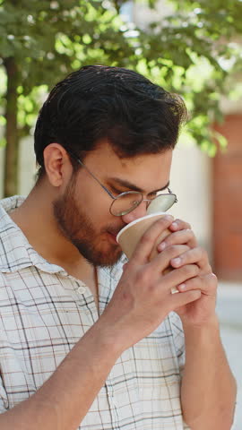 Happy Indian man enjoying morning coffee hot drink and smiling. Relaxing, taking a break. Hispanic young guy standing on urban city center street, drinking coffee to go. Town lifestyles outside.