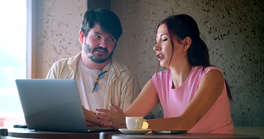 A young man and a young woman, both business professionals, are happily working on their laptops and chatting about work at a café outside the office on a summer day