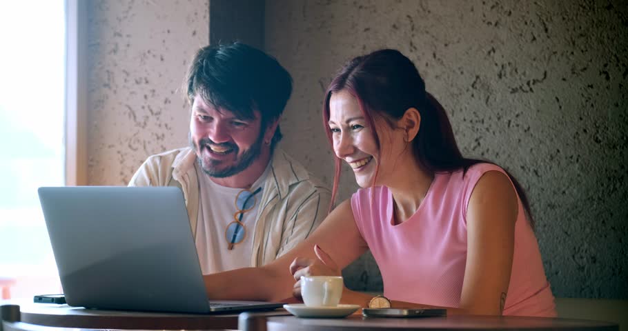 A young man and a young woman, both business professionals, are happily working on their laptops and chatting about work at a café outside the office on a summer day