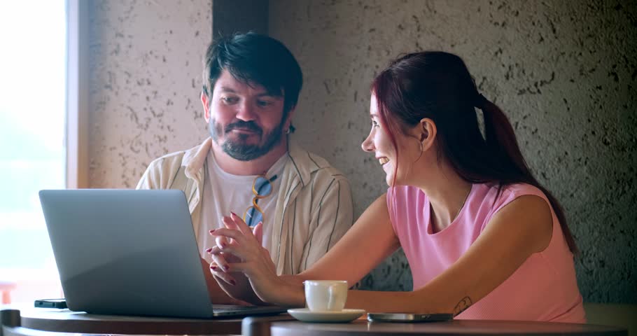 A young man and a young woman, both business professionals, are happily working on their laptops and chatting about work at a café outside the office on a summer day