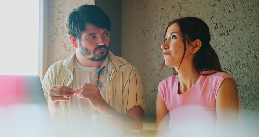 A young man and a young woman, both business professionals, are happily working on their laptops and chatting about work at a café outside the office on a summer day