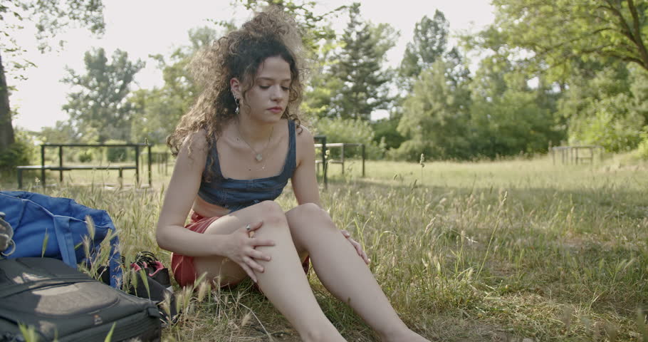 A curly-haired girl takes a break from her modern dance practice, sitting in a meadow within the park, looking relaxed and content.