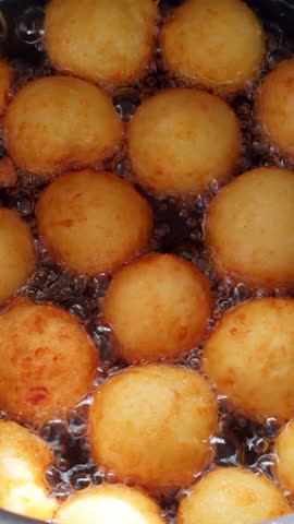 Lots of donuts with no hole in the middle are fried in oil deep. Close up view on the sweet dough pastry cooking. Vertical video.