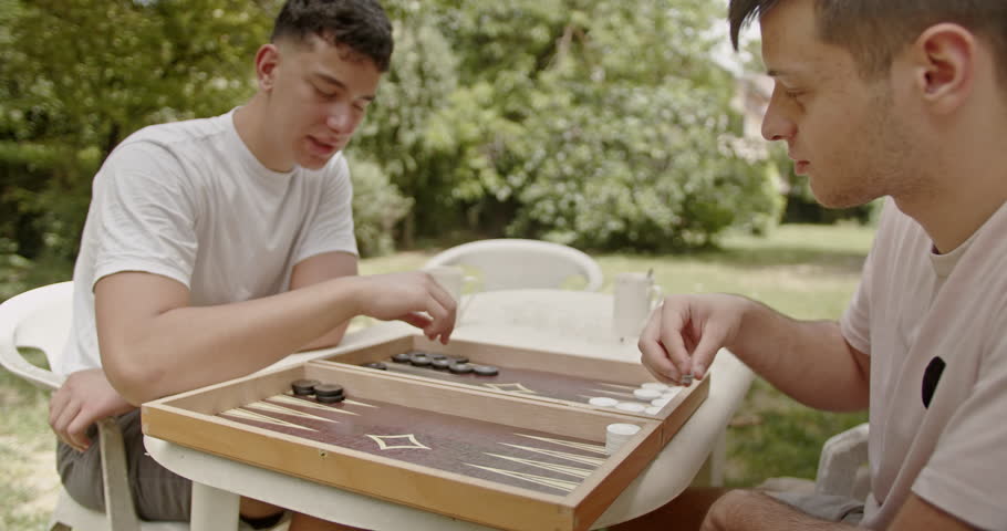 Two young men play a game of backgammon at an outdoor table surrounded by greenery, enjoying the warmth and brightness of a sunny day, immersed in their game and the pleasant surroundings.