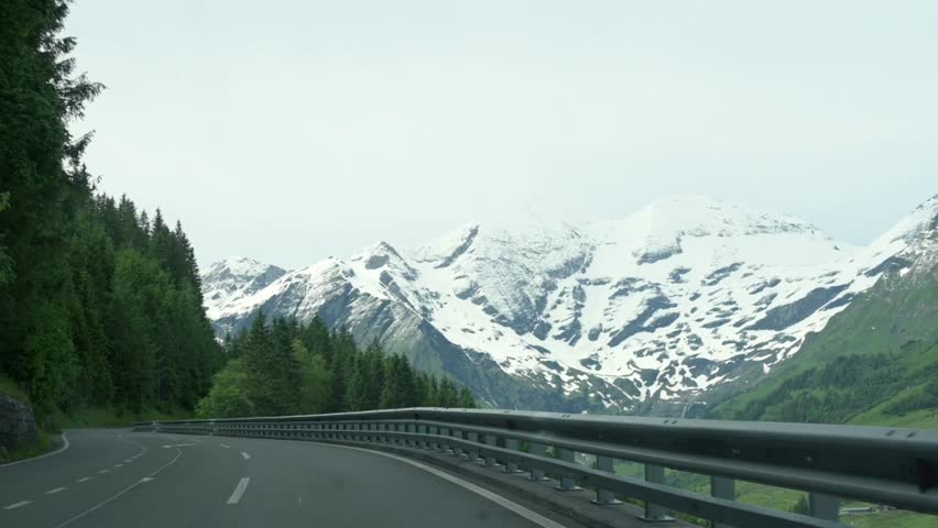 A scenic view of a winding mountain road cutting through a dense forest, with snow-capped peaks in the background at Grossglockner, Austria, highlighting the beauty of an alpine drive.