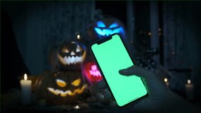 Phone in hand with a green screen against the background of a pile of lanterns glowing with multi-colored lights carved from pumpkins during the celebration of Halloween - Powered by Shutterstock - Get 15% off with code: PIKWIZARD15