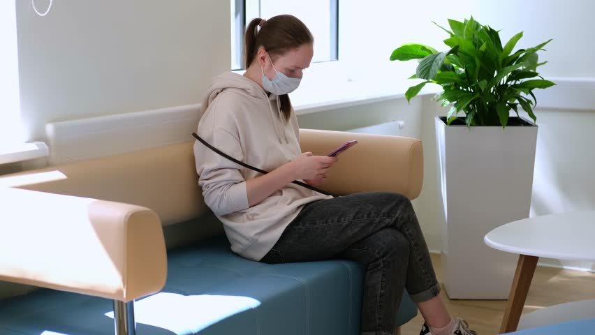 A young white girl wearing a medical mask is using a smartphone in a modern bright interior.