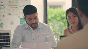 A businessman appears contemplative during a discussion about renewable energy, with models of wind turbines and solar panels in a modern office setting. - Powered by Shutterstock - Get 15% off with code: PIKWIZARD15