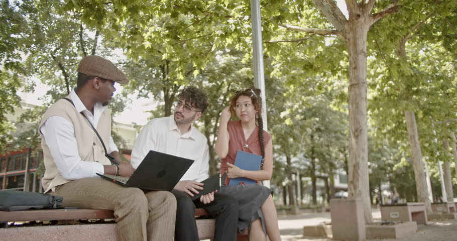 Three colleagues are having an outdoor meeting in a shaded area on a sunny day, working with laptops and discussing project ideas. The relaxed setting enhances their collaboration and brainstorming