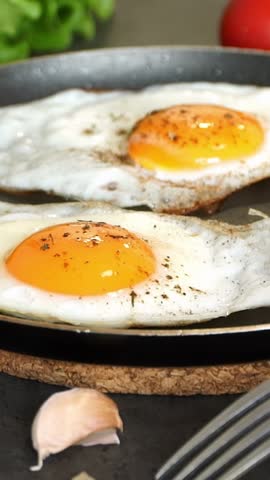 Close up of fried eggs in hot frying pan are sprinkled with spices and seasonings on background of fresh vegetables. Cooking food. Vertical shot
