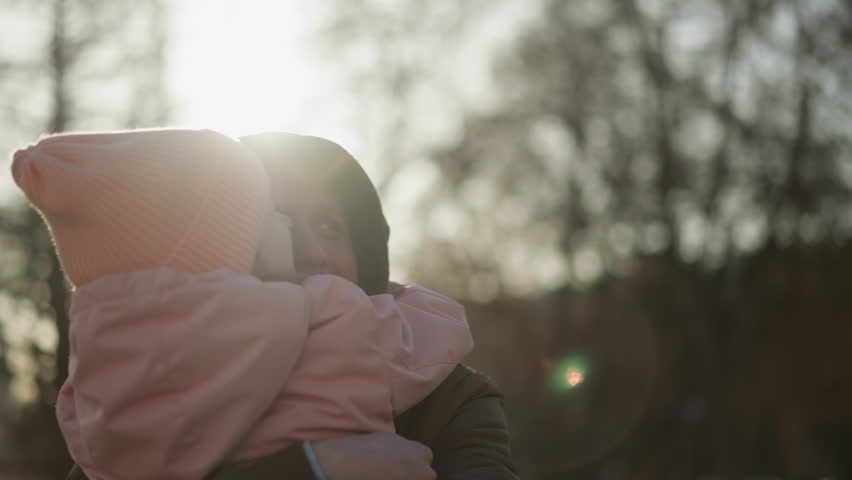 A little girl in a pink cap and jacket being carried by a man wearing a brown jacket, as she lovingly kisses him on the cheek