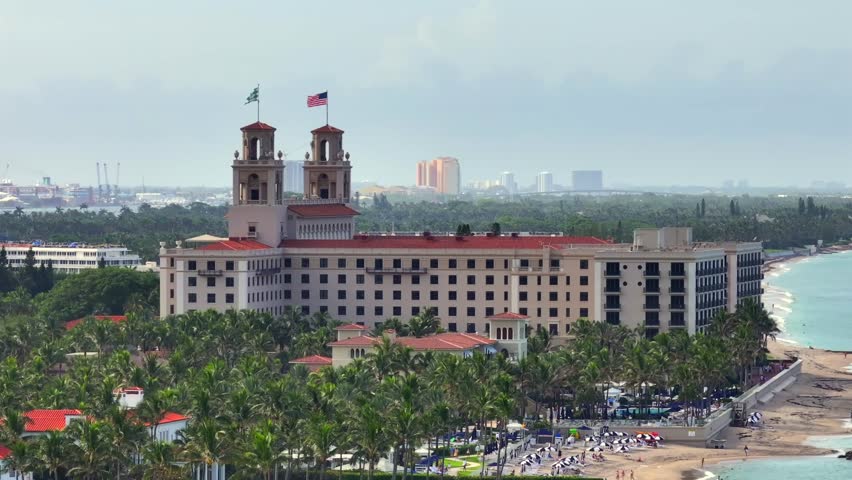 The Breakers Palm Beach resort with beachfront view, surrounded by lush palm trees and iconic twin towers. Oceanfront setting with distant city skyline. Aerial.