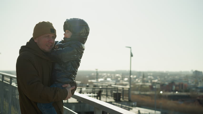 A father stands close to a metal railing, carrying his son who gently places his hand on the father s shoulder, gazing out over a cityscape in the distance