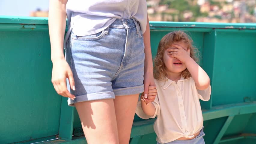 Three-year-old baby is scared and covers her eyes with her hand, her mother stands next to her, they are traveling on cruise ship on sunny summer day, close-up.