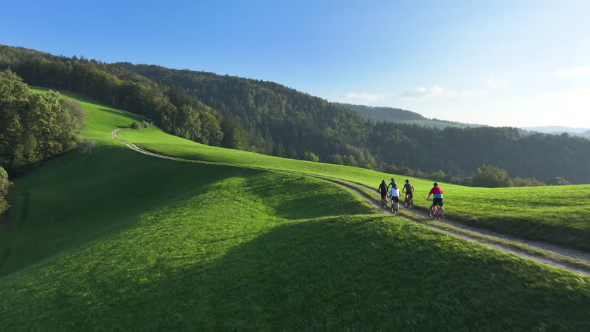 AERIAL: Cycling friends enjoying a serene ride along a green hilltop bike trail bathed in gentle morning light with stunning views of rolling hills and nature. Mountain biking on a sunny autumn day.
